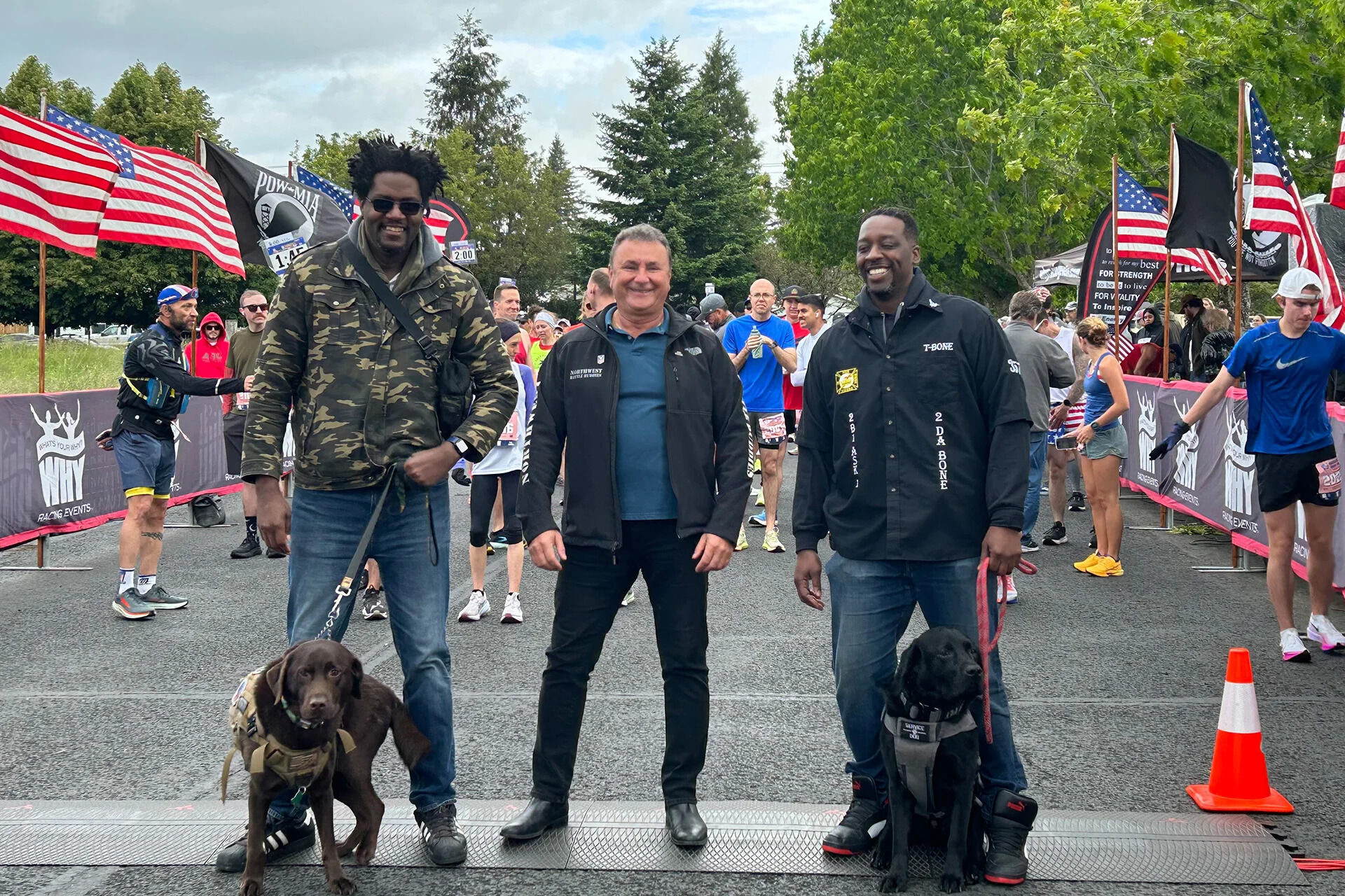 Three men standing next to each other and smiling in front of the starting line of the Reflection Run. Two have service dogs.